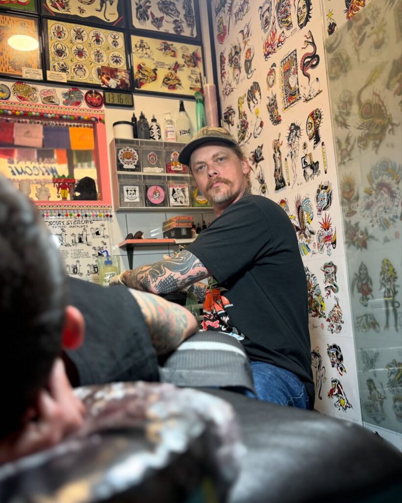 Tatuador trabajando en Hidden Doorway Tattoo, en Ciutat Vella (Barcelona). Estudio lleno de láminas de tatuajes tradicionales en la pared.