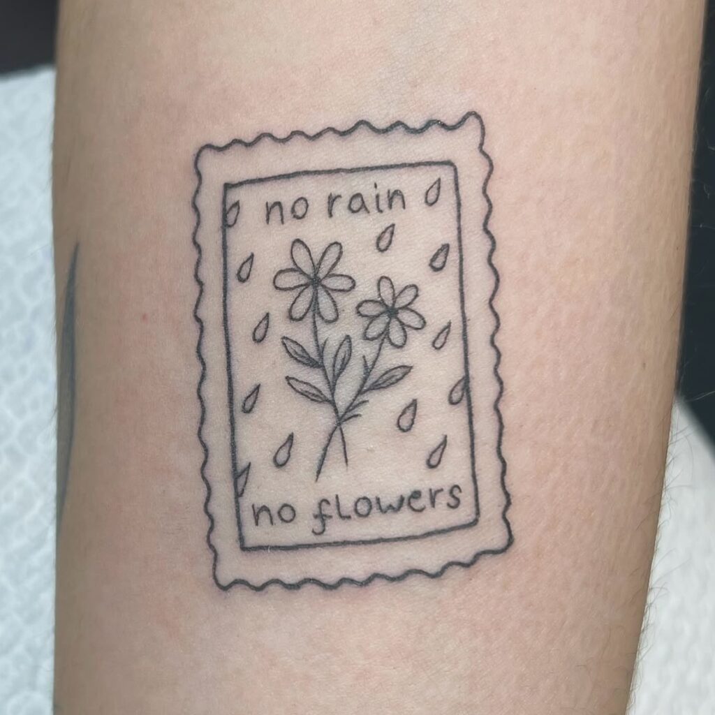 Tatuaje lineal en estilo fino con marco en forma de sello, dos flores y gotas de lluvia con la frase no rain, no flowers, hecho en Barcelona.