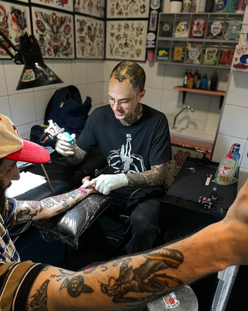 Tatuador trabajando en un estudio de Barcelona, especializado en tatuajes tradicionales y realistas, aplicando tinta en el brazo de un cliente rodeado de diseños coloridos en la pared.
