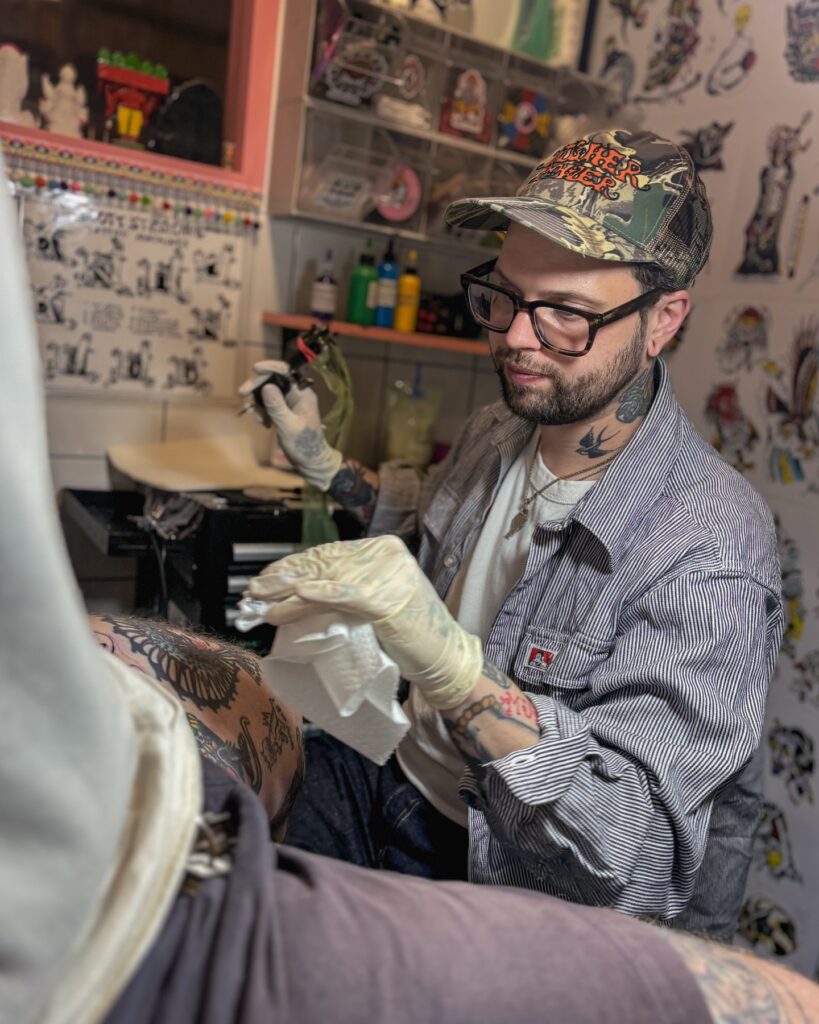 Tatuador trabajando en un estudio de tatuajes en Barcelona, usando guantes y máquina de tatuar mientras realiza un diseño en la pierna de un cliente. En la pared del estudio se ven láminas con dibujos y diseños de tatuajes.